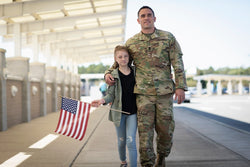 Military father dressed in army fatigues walking down airport terminal with daughter holding American flag.